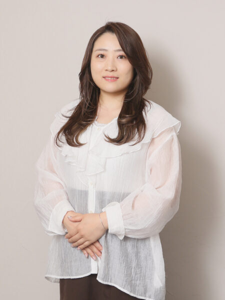 Portrait of a woman with brown hair wearing a white sheer blouse, standing against a light gray wall.