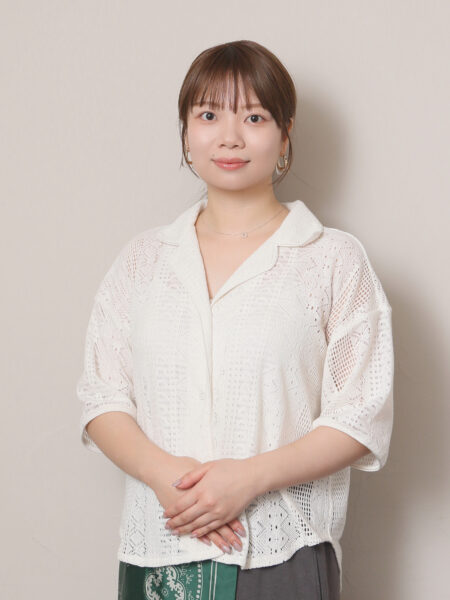 Young woman standing against a beige background, wearing a white lace cardigan and a green patterned skirt, with hands gently clasped in front.