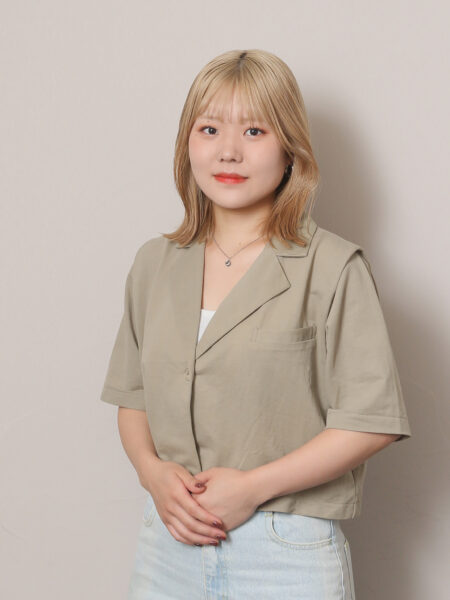 Portrait of a young woman with shoulder-length blonde hair, wearing a beige blazer and light jeans, standing against a neutral backdrop.