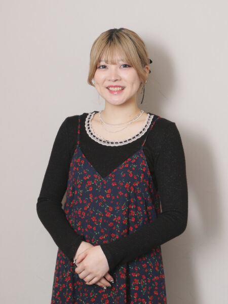 Smiling young woman with blonde hair wearing a navy floral dress over a black top and layered necklaces, standing against a light gray background.